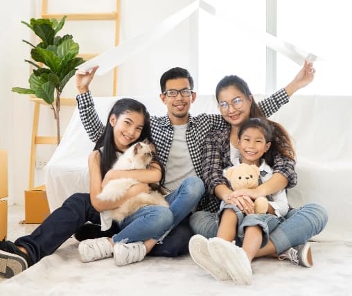 Happy family posing together in a cozy, light-filled living space: a father in glasses and a checked shirt sits in the middle with a joyful expression, alongside a mother in a black and white patterned shirt, both smiling warmly. A teenage girl on one side holds a fluffy cat in a casual outfit, while a younger boy on the other side sits with a teddy bear, looking cheerful and relaxed. The background features a light-colored sofa, a decorative plant, and soft natural light from a window, creating a warm and inviting atmosphere. The close grouping and relaxed poses convey a strong sense of family love and happiness in a comfortable home environment.